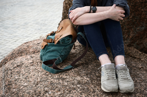 A close shot of the legs of a girl sitting on a stone and a backpack nearby.