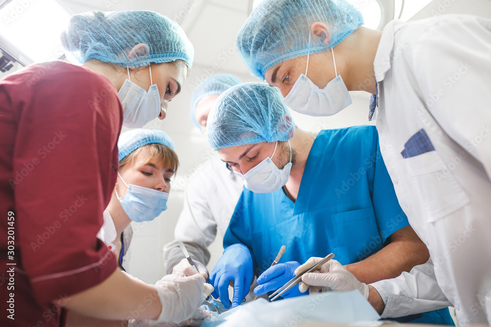 Group of surgeons at work operating in surgical theatre. Resuscitation medicine team wearing protective masks holding steel medical tools saving patient. Surgery and emergency concept. Group of surgeons at work operating in surgical theatre. Resuscitation medicine team wearing protective masks holding steel medical tools saving patient. Surgery and emergency concept.