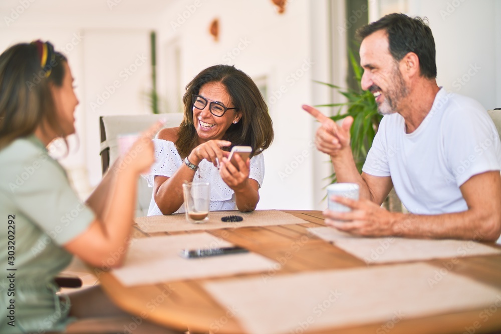 Beautiful family sitting on terrace drinking cup of coffee using smartphone speaking and smiling