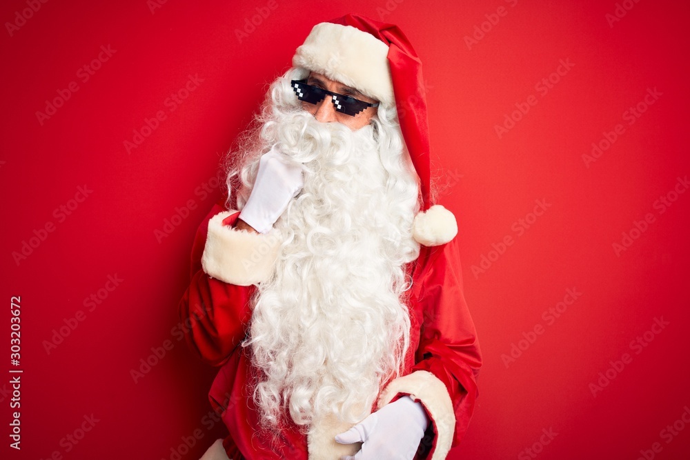 Middle age man wearing Santa Claus costume and sunglasses over isolated red background with hand on chin thinking about question, pensive expression. Smiling with thoughtful face. Doubt concept.