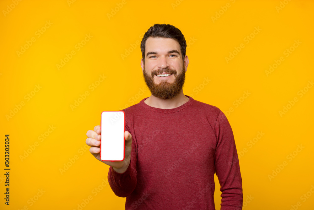 Photo of cheerful guy with beard and wearing red sweater, showing mobile screen at camera over yellow background Photo of cheerful guy with beard and wearing red sweater, showing mobile screen at camera over yellow background