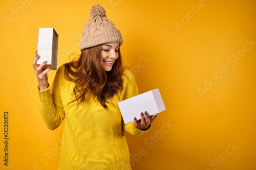 The brunette stands on a yellow background in a yellow sweater and hat, and smiles and looks into the gift box, lifting the lid. photo