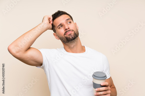 Young man with beard holding a take away coffee over isolated blue background having doubts and with confuse face expression