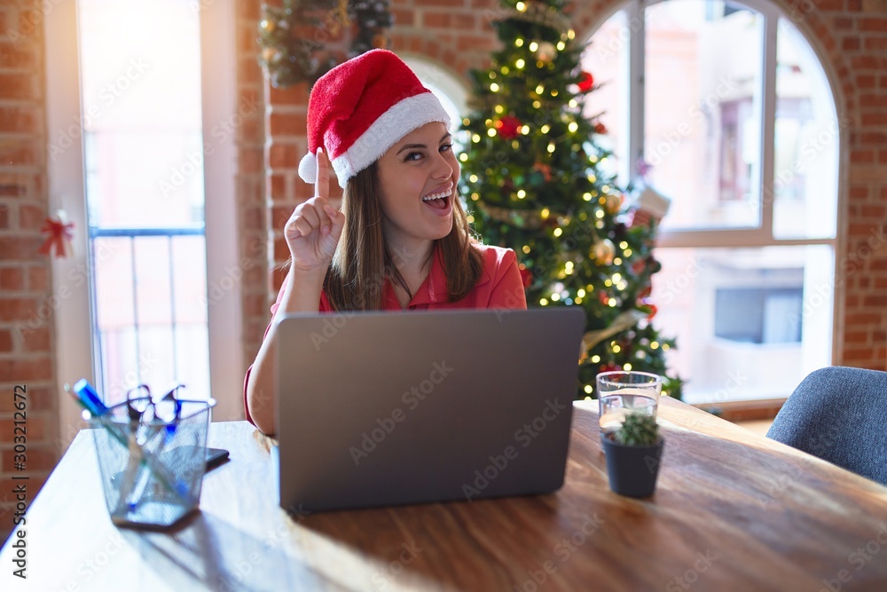 Beautiful woman sitting at the table working with laptop wearing santa claus hat at christmas pointing finger up with successful idea. Exited and happy. Number one. Beautiful woman sitting at the table working with laptop wearing santa claus hat at christmas pointing finger up with successful idea. Exited and happy. Number one.