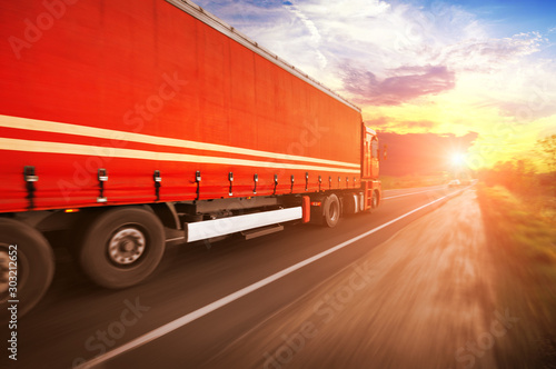 Truck with a trailer driving fast on the countryside road against a sky with a sunset