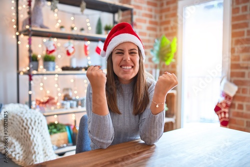 Young beautiful woman wearing christmas hat sitting at the table at home celebrating surprised and amazed for success with arms raised and open eyes. Winner concept.