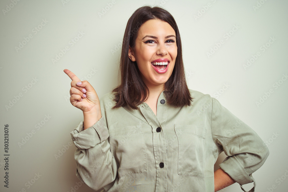 Young beautiful brunette woman wearing green shirt over isolated background with a big smile on face, pointing with hand and finger to the side looking at the camera. Young beautiful brunette woman wearing green shirt over isolated background with a big smile on face, pointing with hand and finger to the side looking at the camera.