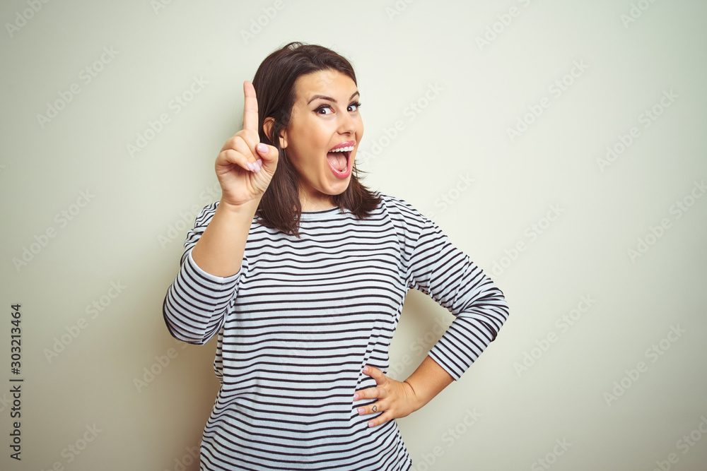 Young beautiful brunette woman wearing striped sweater over white isolated background pointing finger up with successful idea. Exited and happy. Number one. Young beautiful brunette woman wearing striped sweater over white isolated background pointing finger up with successful idea. Exited and happy. Number one.