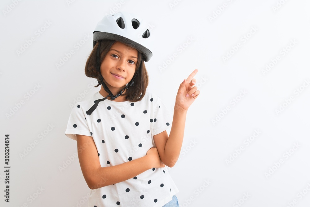 Beautiful child girl wearing security bike helmet standing over isolated white background with a big smile on face, pointing with hand and finger to the side looking at the camera. Beautiful child girl wearing security bike helmet standing over isolated white background with a big smile on face, pointing with hand and finger to the side looking at the camera.
