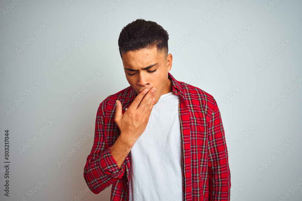 Young brazilian man wearing red shirt standing over isolated white background bored yawning tired covering mouth with hand. Restless and sleepiness.