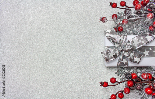 Bożonarodzeniowe srebrne tło z prezentem, gałązkami pokrytymi czerwonymi jagodami oraz srebrnymi gwiazdkami