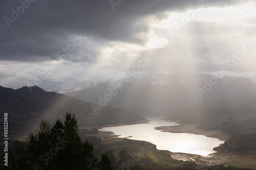 The large reservoir Tranco in the Spanish mountains of Andalusia. The sky is very cloudy and broad sunbeams illuminate the landscape and the lake. It s sunset soon.