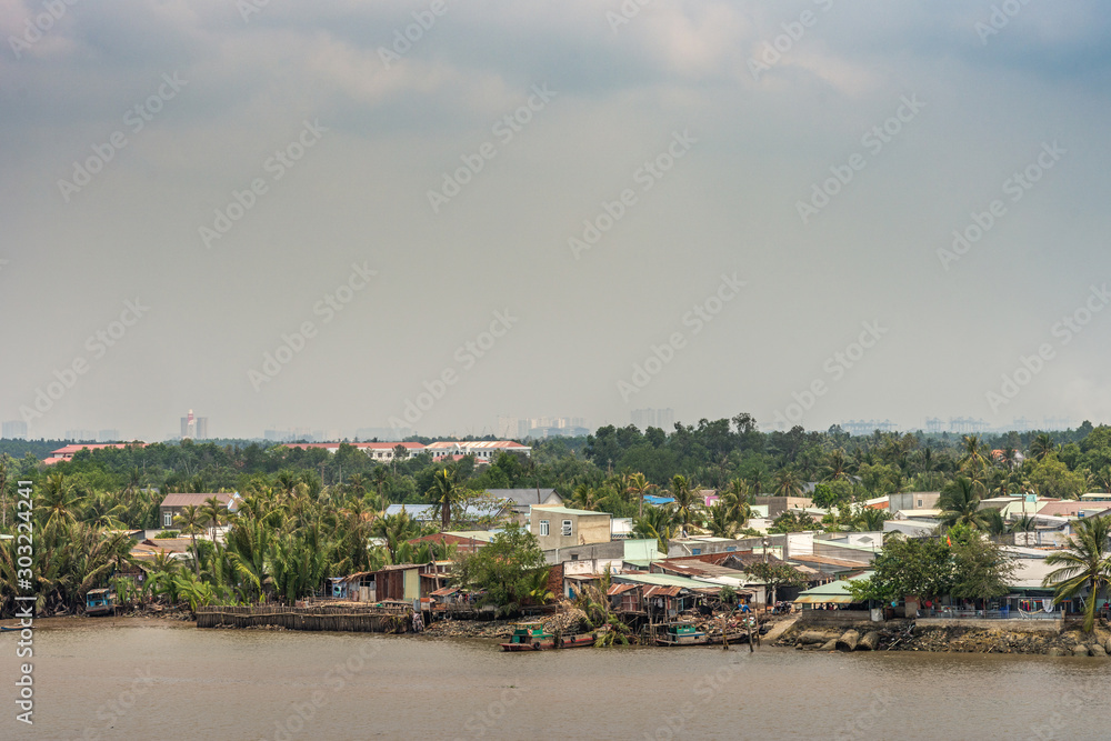 Long Tau River, Vietnam - March 12, 2019: Riverside Phuoc Khanh village with multiple houses. Light blue cloudscape and green vegetation along shore. In distance, high rise buildings of Ho Chi Minh Ci