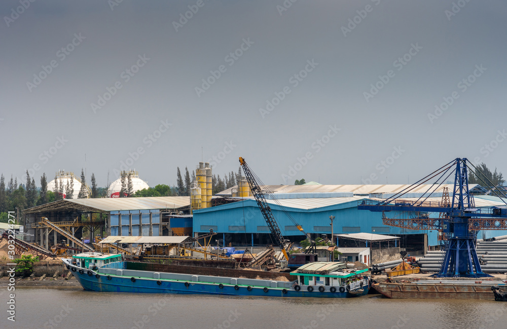 Long Tau River, Vietnam - March 12, 2019: River cargo boat docked on quay in front of blue atelier, warehouse, and being worked with black crane under light blue sky. Chaotic scenery.