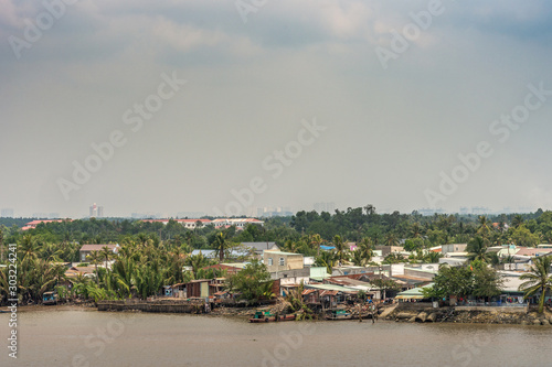 Long Tau River, Vietnam - March 12, 2019: Riverside Phuoc Khanh village with multiple houses. Light blue cloudscape and green vegetation along shore. In distance, high rise buildings of Ho Chi Minh Ci © Klodien
