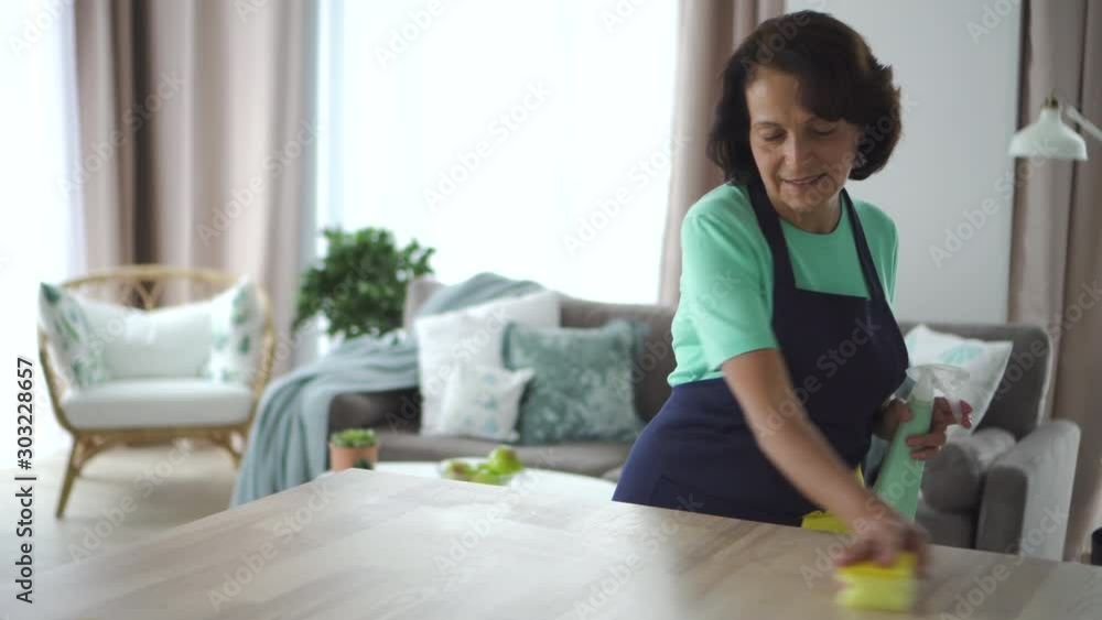 60s Happy american mature housewife clean and polish table in living room. 4k Beautiful elderly female wash desk, remove dust, holding spray bottle in hand. Smiling 60s brunette doing housework 60s Happy american mature housewife clean and polish table in living room. 4k Beautiful elderly female wash desk, remove dust, holding spray bottle in hand. Smiling 60s brunette doing housework
