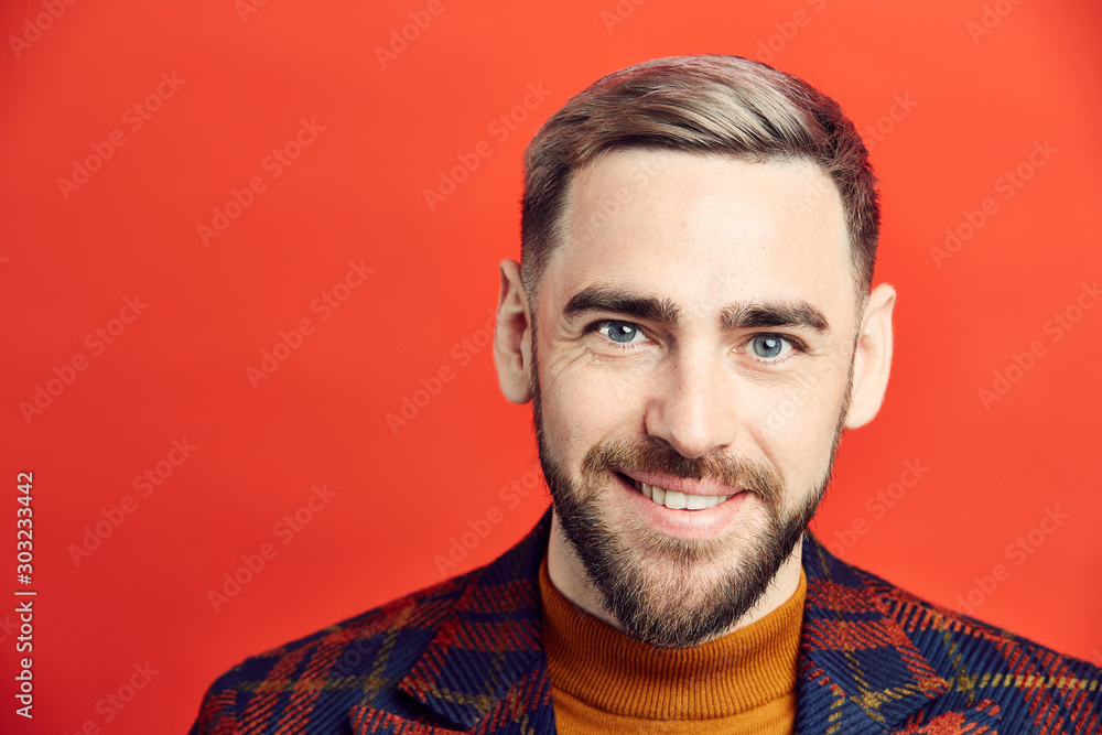 Obraz premium Head and shoulders portrait of smiling bearded man looking at camera while posing against red background, copy space