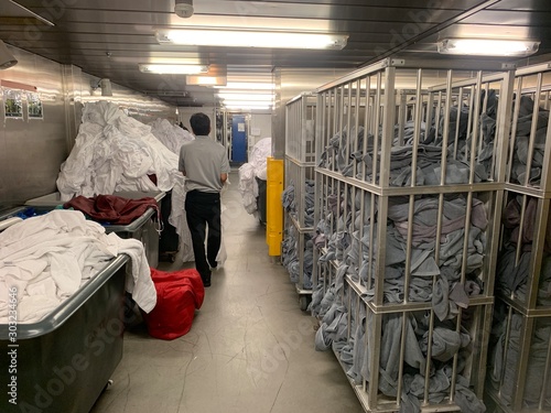 Cleaner is walking in yhr huge laundry on a cruise ship. A lot of white dirty linen and grey pool towels in the trolley.