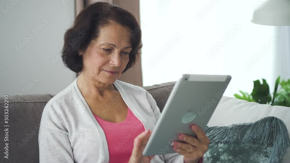 Happy american businesswoman using tablet sitting at sofa in living room. Smiling mature female browsing web and having shopping online, holding gadget in hand. Portrait of old lady is relaxing at Happy american businesswoman using tablet sitting at sofa in living room. Smiling mature female browsing web and having shopping online, holding gadget in hand. Portrait of old lady is relaxing at