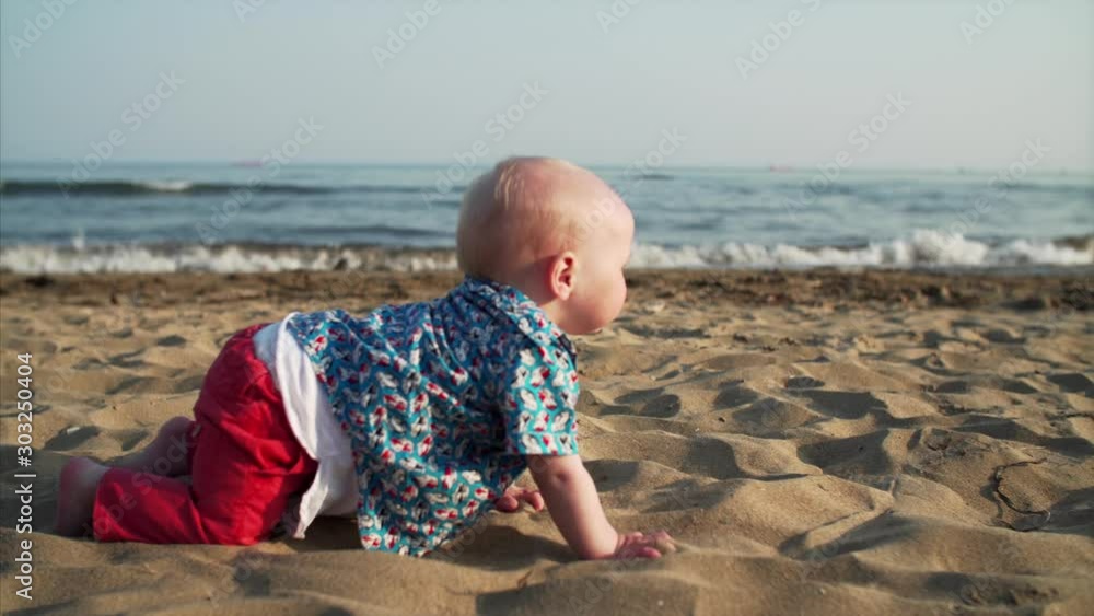 Adorable caucasian baby boy is crawling on sandy beach at seaside. Sea is on the background. Sunny weather Adorable caucasian baby boy is crawling on sandy beach at seaside. Sea is on the background. Sunny weather