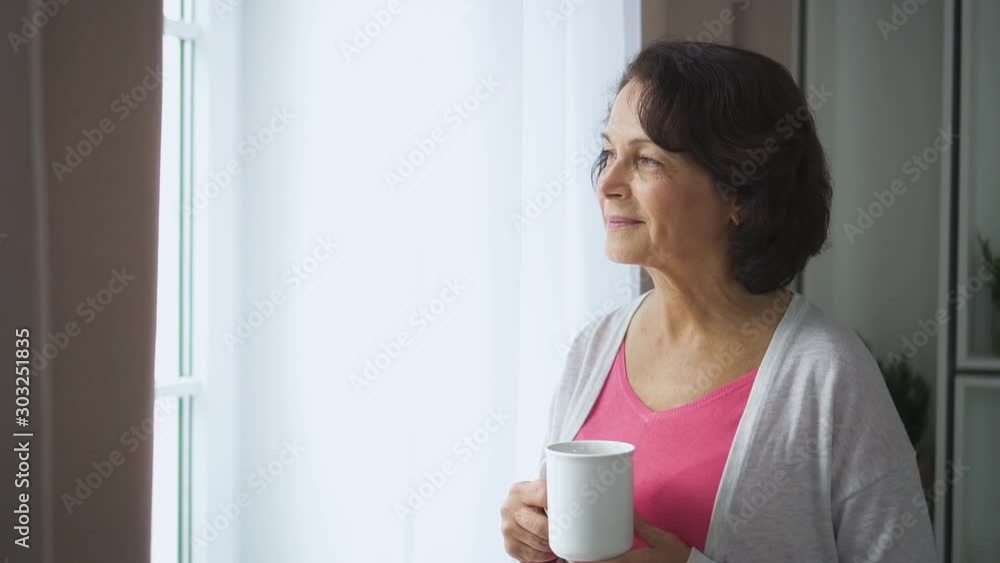 American elderly woman drinking coffee and watching window in home room. Smiling mature female holding mug in hand, drink beverage standing in apartment interior. Elegant 60s lady wearing casual American elderly woman drinking coffee and watching window in home room. Smiling mature female holding mug in hand, drink beverage standing in apartment interior. Elegant 60s lady wearing casual