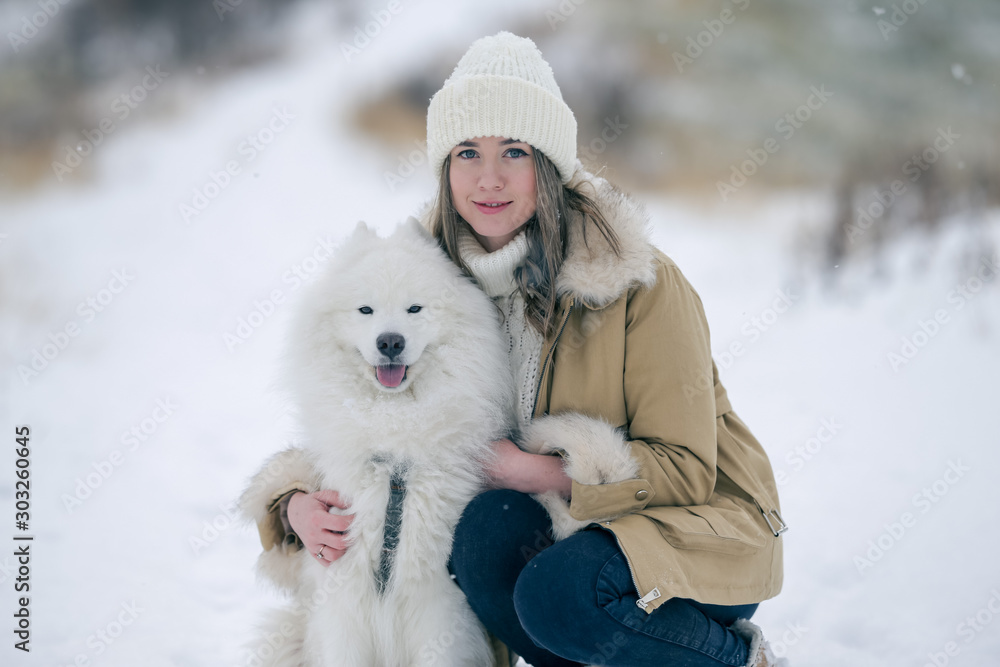 A young girl walks in winter with a white Samoyed dog in a snowy meadow in the forest. A young girl walks in winter with a white Samoyed dog in a snowy meadow in the forest.