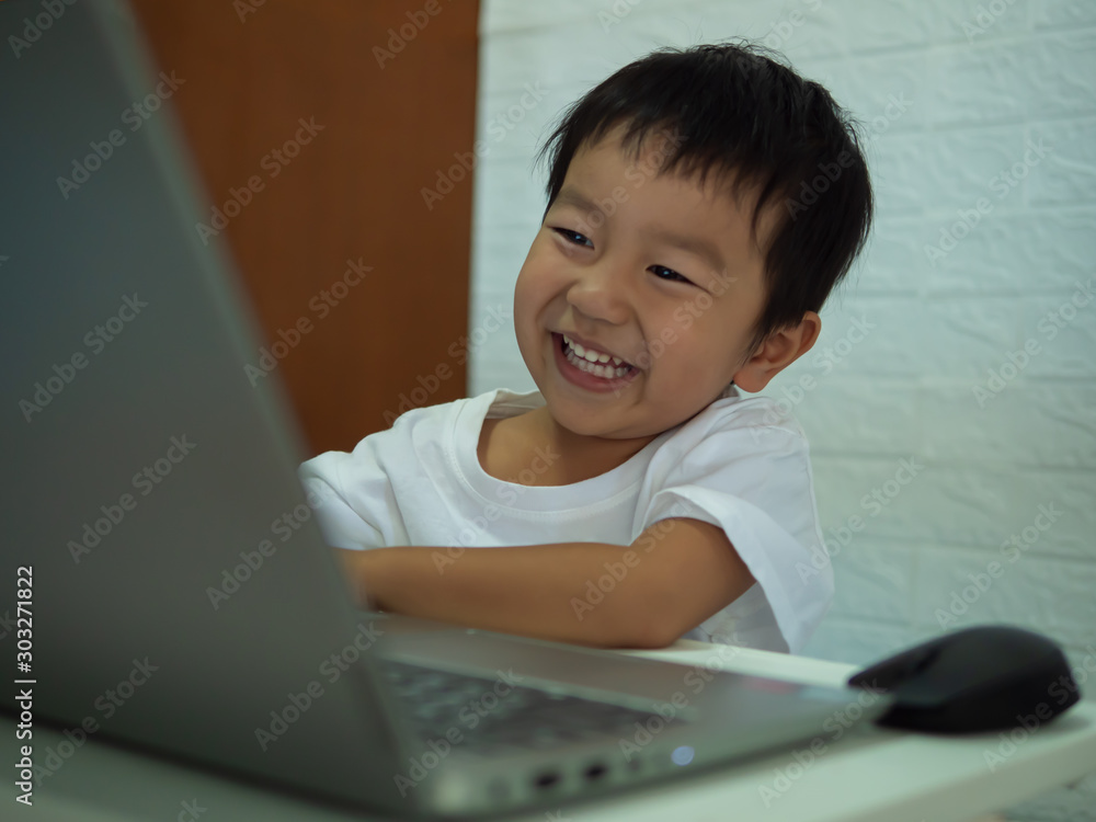 Asian child boy using notebook or computer for learning. Kid in white t-shirt with laughing and smiling on face looking to laptop to search internet at home.