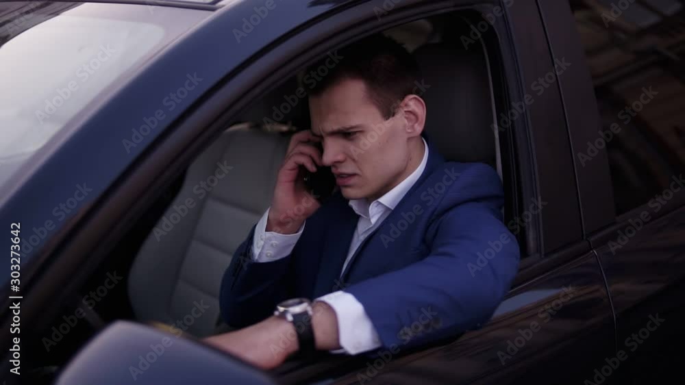 Handsome man sitting in the car on a driver's seat, holding hand on the car door. Businessman talking while sitting in a car. Serious man in blue suit using a mobile phone. Slow motion Handsome man sitting in the car on a driver's seat, holding hand on the car door. Businessman talking while sitting in a car. Serious man in blue suit using a mobile phone. Slow motion