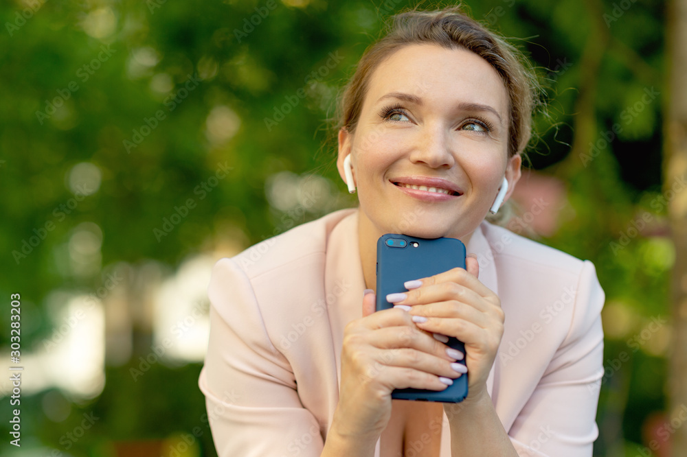Close-up portrait of a smiling woman with smartphone on the street. Happy businesswoman is using phone, outdoors. Cheerful businesswoman in a jacket with cell phone in park looking away. Close-up portrait of a smiling woman with smartphone on the street. Happy businesswoman is using phone, outdoors. Cheerful businesswoman in a jacket with cell phone in park looking away.