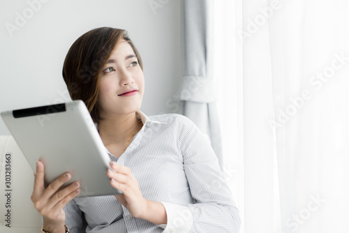 Portrait of a happy Asian woman sitting on sofa using Digital Tablet near window at home.