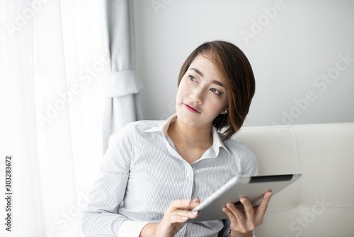Portrait of a happy Asian woman sitting on sofa using Digital Tablet near window at home.