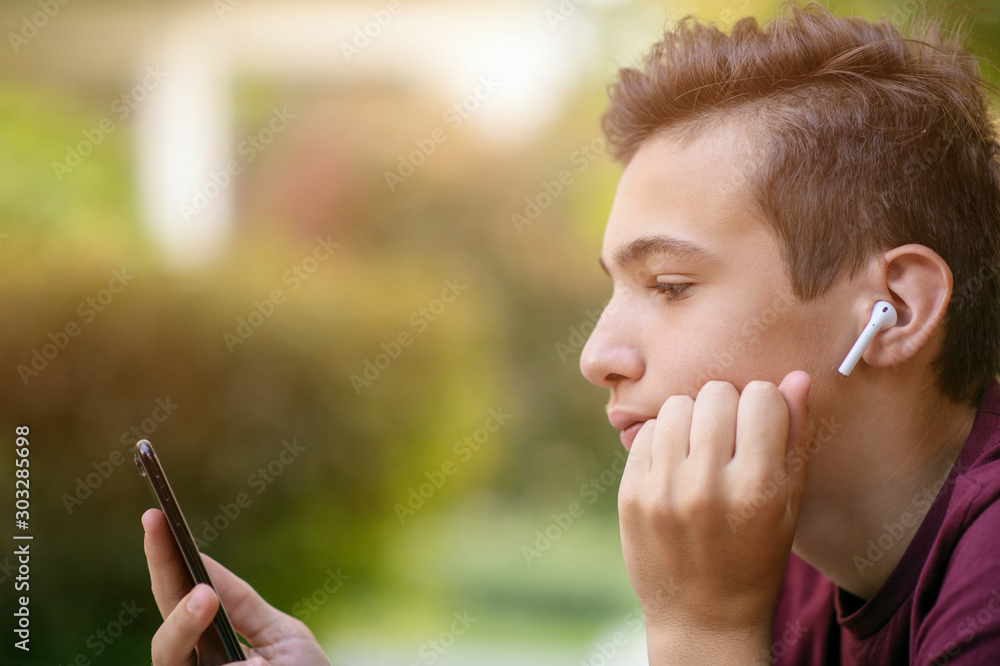 Close-up portrait of a thoughtful unhappy teenage boy with smartphone, outdoors.  Sad teenager with mobile phone looks away, in the park.  Pensive teenager in casual clothes with cell phone in park - obrazy, fototapety, plakaty 