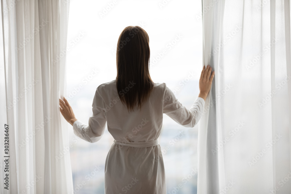 Young woman open curtain looking outside window, rear back view Young woman open curtain looking outside window, rear back view