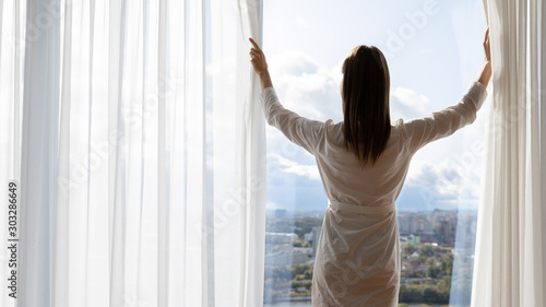 Rich young woman wear gown open curtains stand at window