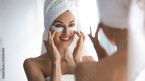 Happy woman towel on head apply patches looking in mirror photo