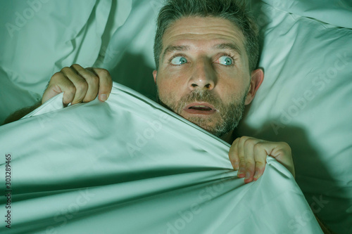  stressed and scared man alone in bed awake at night in fear after having a nightmare feeling paranoid holding the blanket in funny panic face expression photo