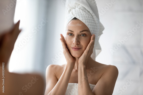 Beautiful young woman look in mirror massaging face applying cream