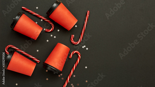 Paper coffee cup with sugar canes on a black background. Christmas background with Xmas candy canes and star confetti. Top view, flat lay, mockup. Copy space for text.