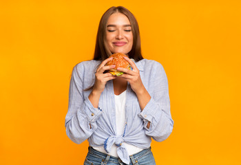 Cheerful Woman Smelling Burger Standing Over Yellow Background