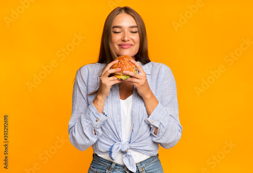 Cheerful Woman Smelling Burger Standing Over Yellow Background