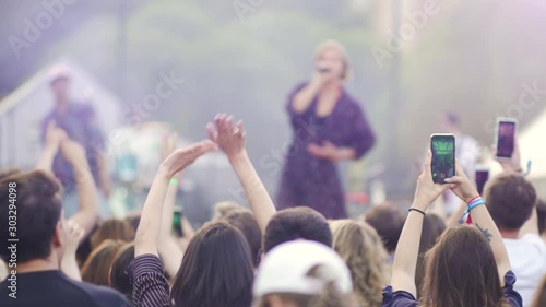 Famous artists performing front of american fans at rock festival in summer day. Young people making photo and recording video holding smartphone in hand at musical concert in green park. Back view of