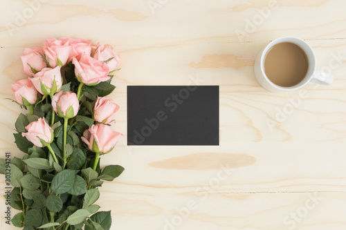 Top view of a black card mockup with a bouquet of pink roses and a coffee on a wooden table.