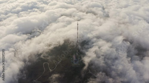 Red and white colored telecommunication tower in dense clouds of fog moving  over the hills in the afternoon. Slow push in aerial shot. photo