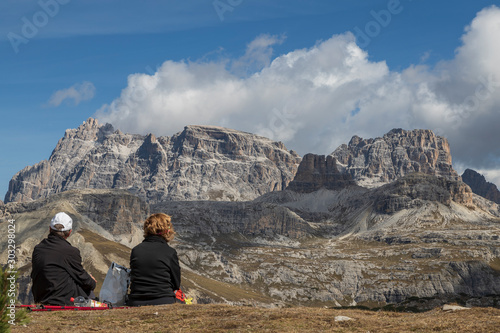 People looking into a valley landscape under Tre Cime in the Italian Dolomites. The weather is nice and there are white clouds in the blue sky.