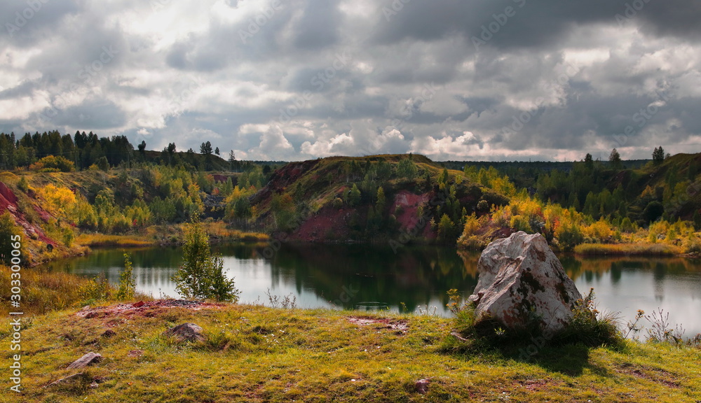 A large stone lying on a cliff by the water. View from the cliff to an abandoned quarry. The quarry is filled with water. Heavy autumn clouds over the reservoir. 