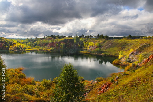 The shores of an abandoned quarry are illuminated by the rays of the sun on a cloudy day through heavy autumn clouds. View from the cliff to an abandoned quarry. The quarry is filled with water.