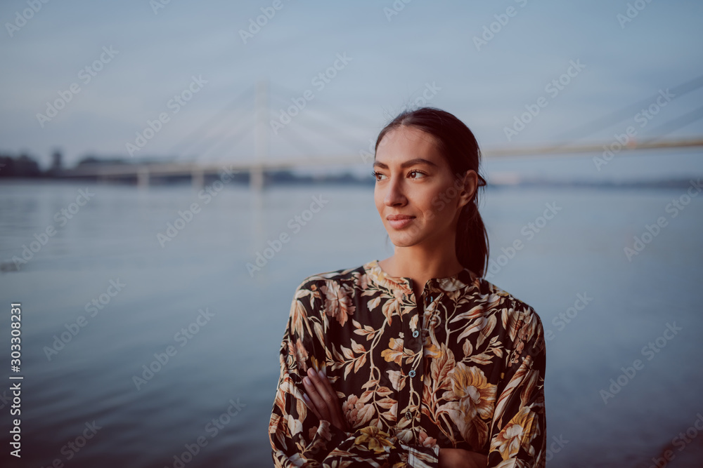 Fototapeta premium Beautiful caucasian brunette in floral dress standing on shore with hands folded and looking away. In background is bridge.