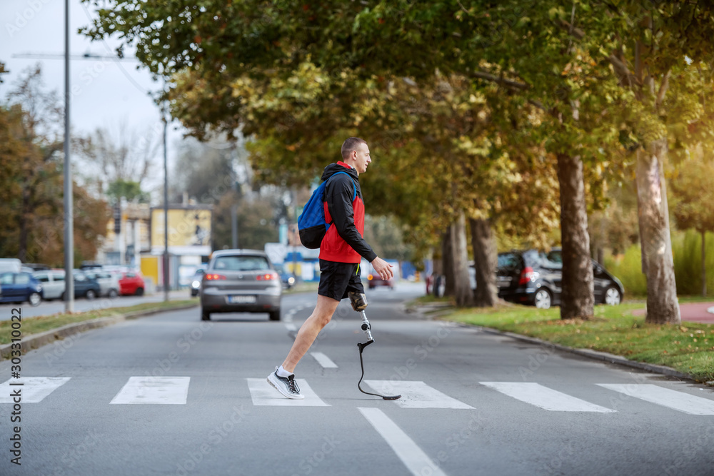 Full length of handsome caucasian handicapped sportsman in sportswear, with artificial leg and backpack crossing street.