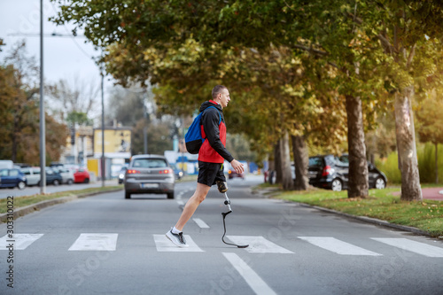 Full length of handsome caucasian handicapped sportsman in sportswear, with artificial leg and backpack crossing street.