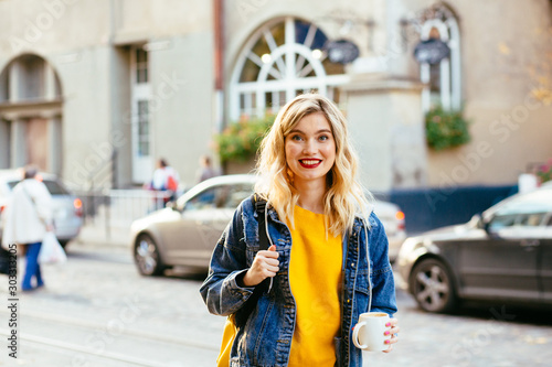 The concept of street fashion. young stylish girl student wearing boyfriend jeans, white sneakers bright yellow sweatshirt.She holds coffee to go. portrait of smiling girl in sunglasses and with bag